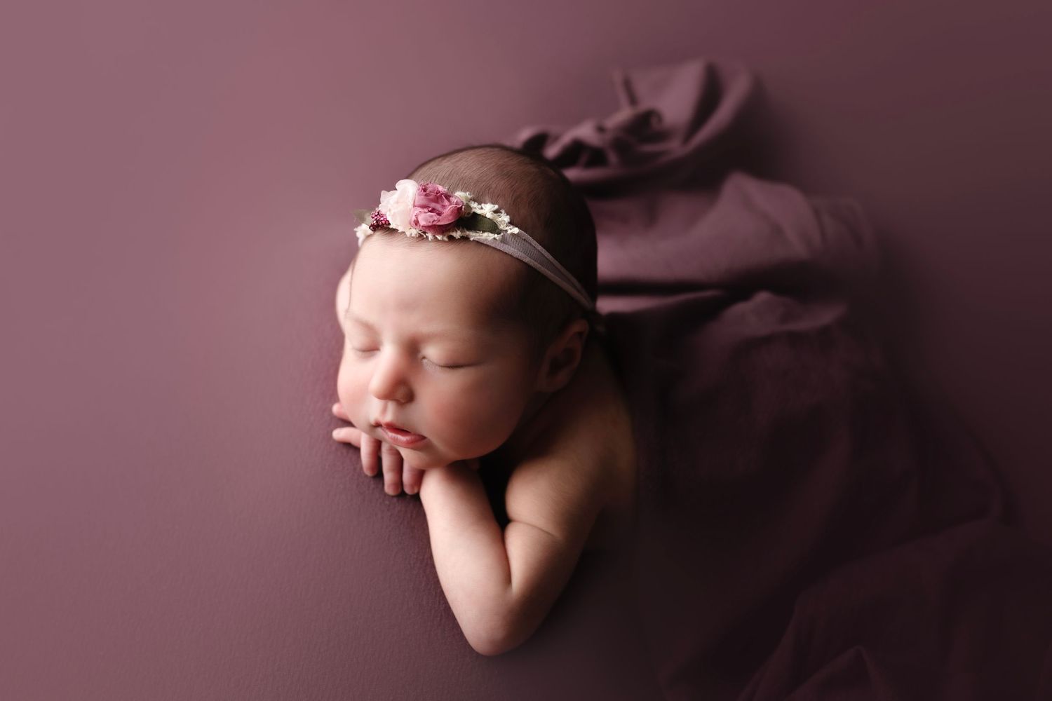 Sleeping newborn baby wearing delicate floral headband rests peacefully on soft mauve backdrop during studio photoshoot.