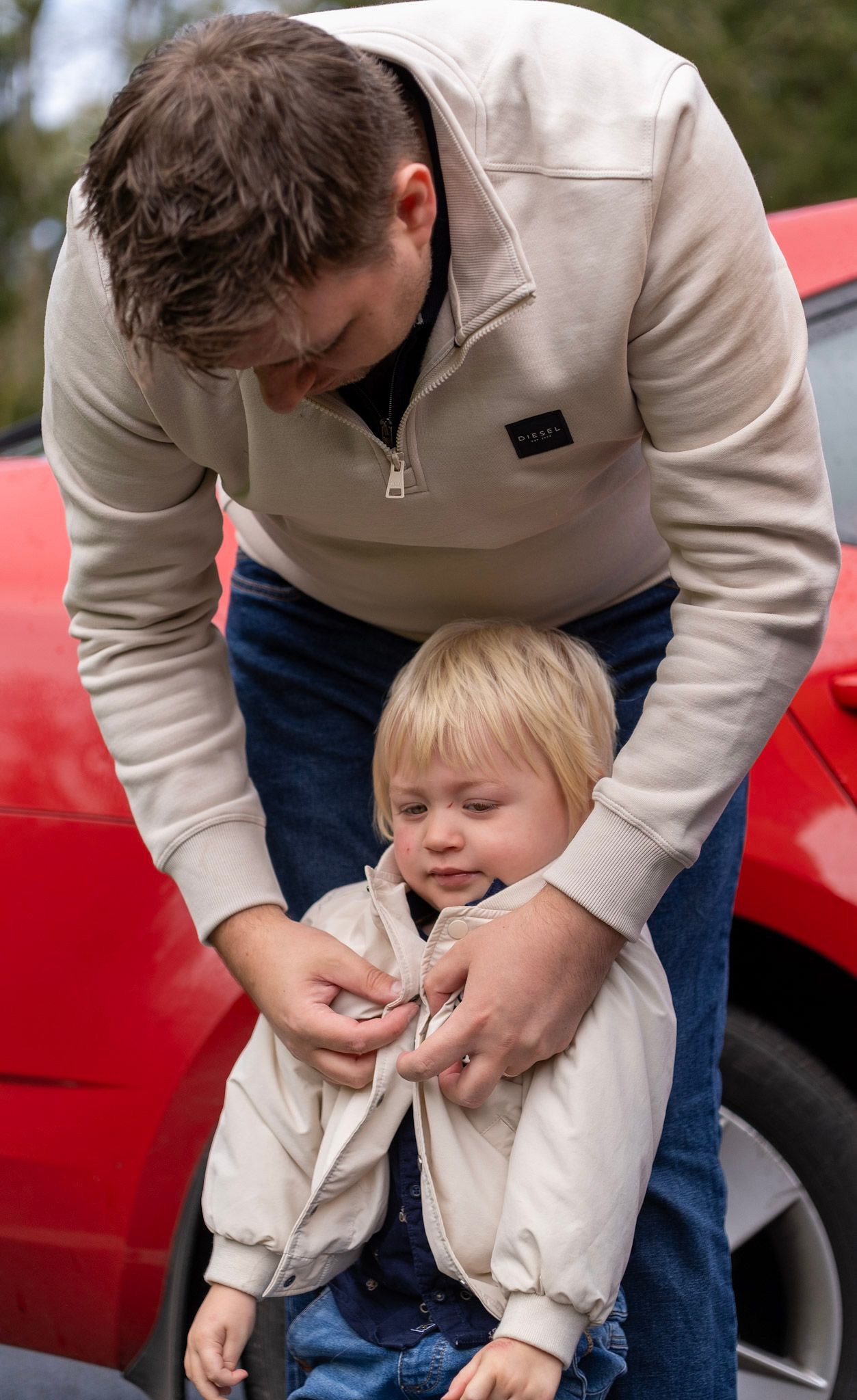Adult helps child with jacket zipper beside red car.