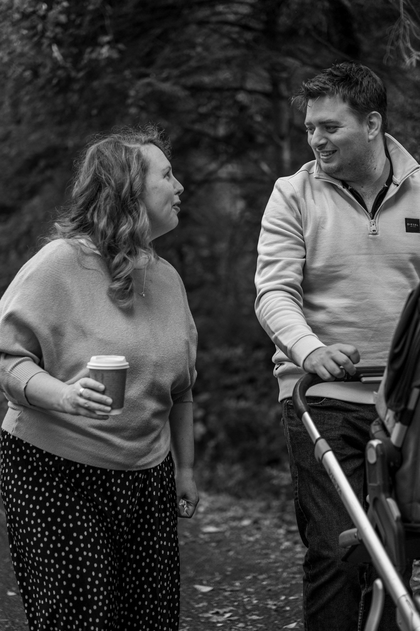 Two people share conversation while holding coffee cups on forest path.