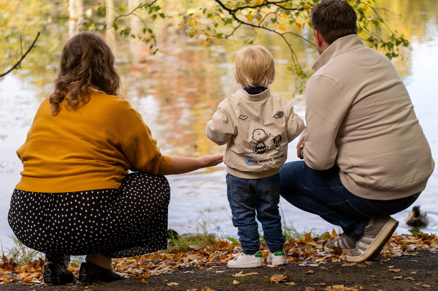 Family sits by water's edge watching autumn leaves fall.