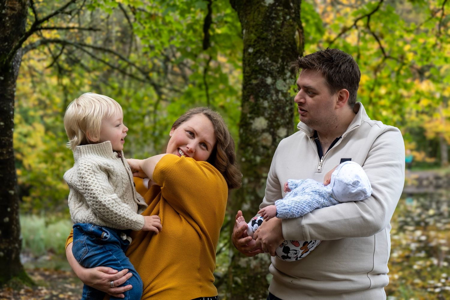 Family of three stands together holding children in autumn forest setting.