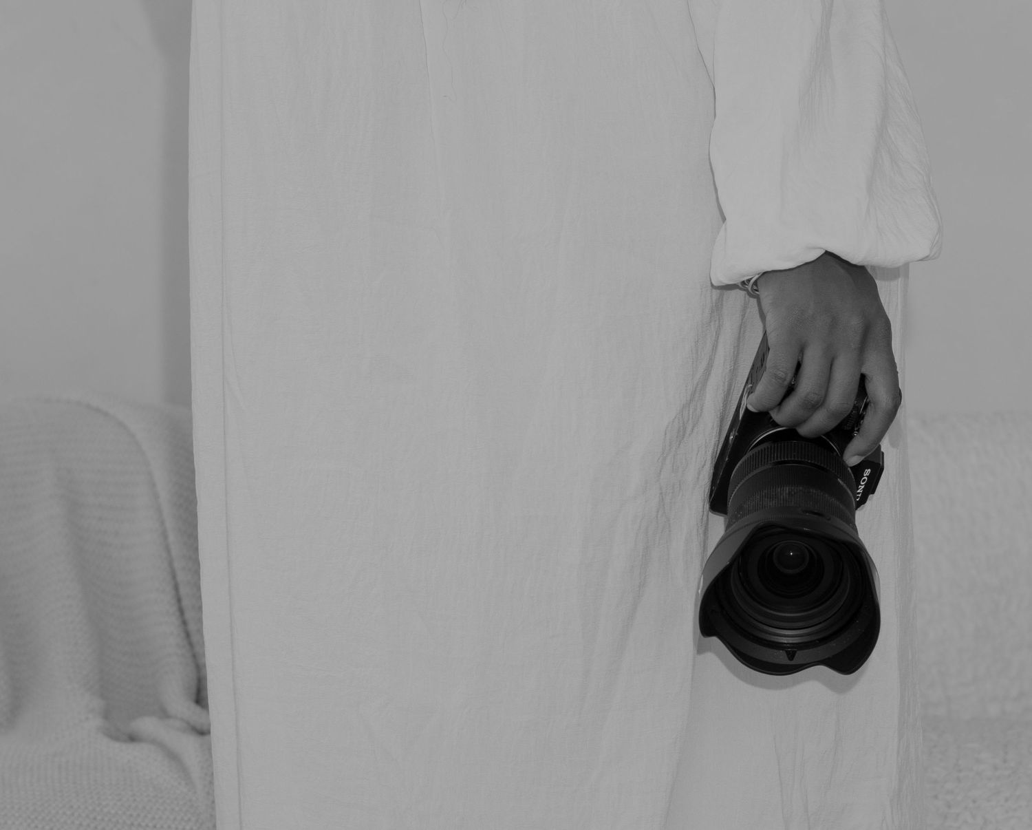 A person in white clothing holds a camera lens at their side in this minimalist black and white photograph.