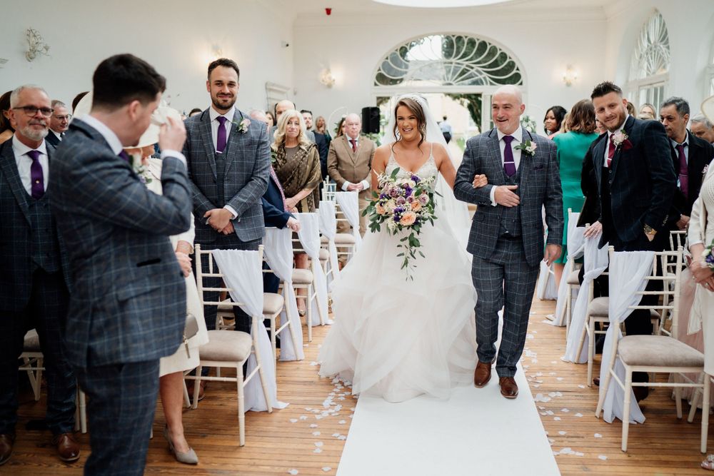 Couple walking down white aisle during wedding ceremony while guests look on.