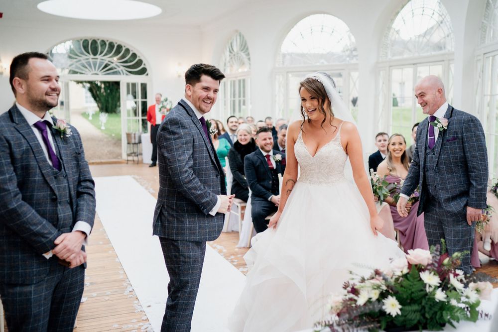 Wedding ceremony in bright indoor venue with elegant decor and white chairs.