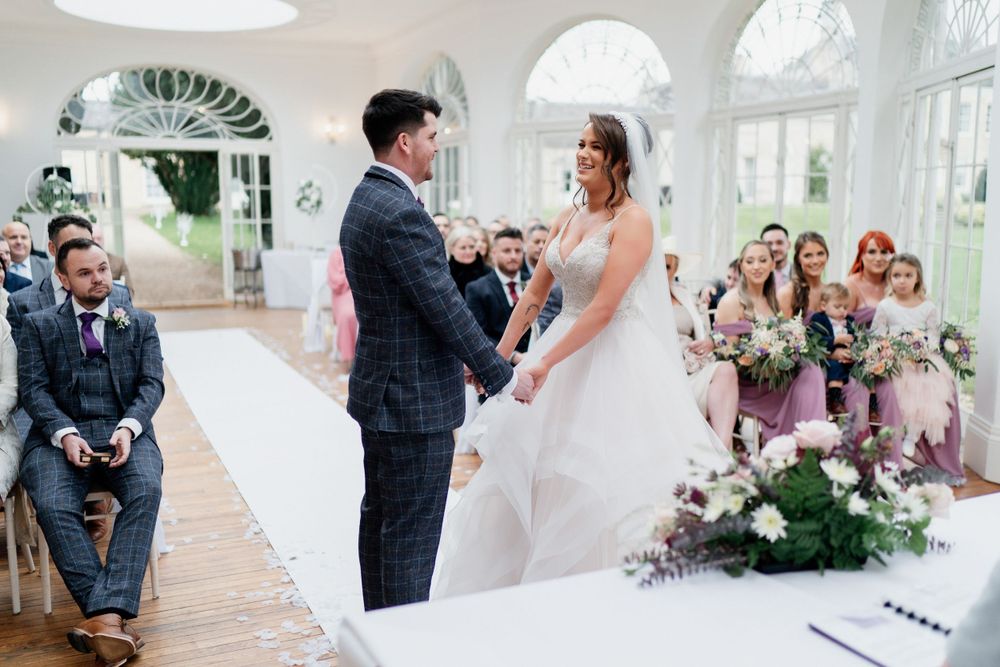 Wedding ceremony with floral decorations in bright indoor venue with arched windows.