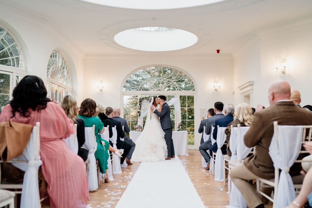Wedding ceremony at Barton Hall in a bright sunlit room with arched windows and guests seated in white chairs.