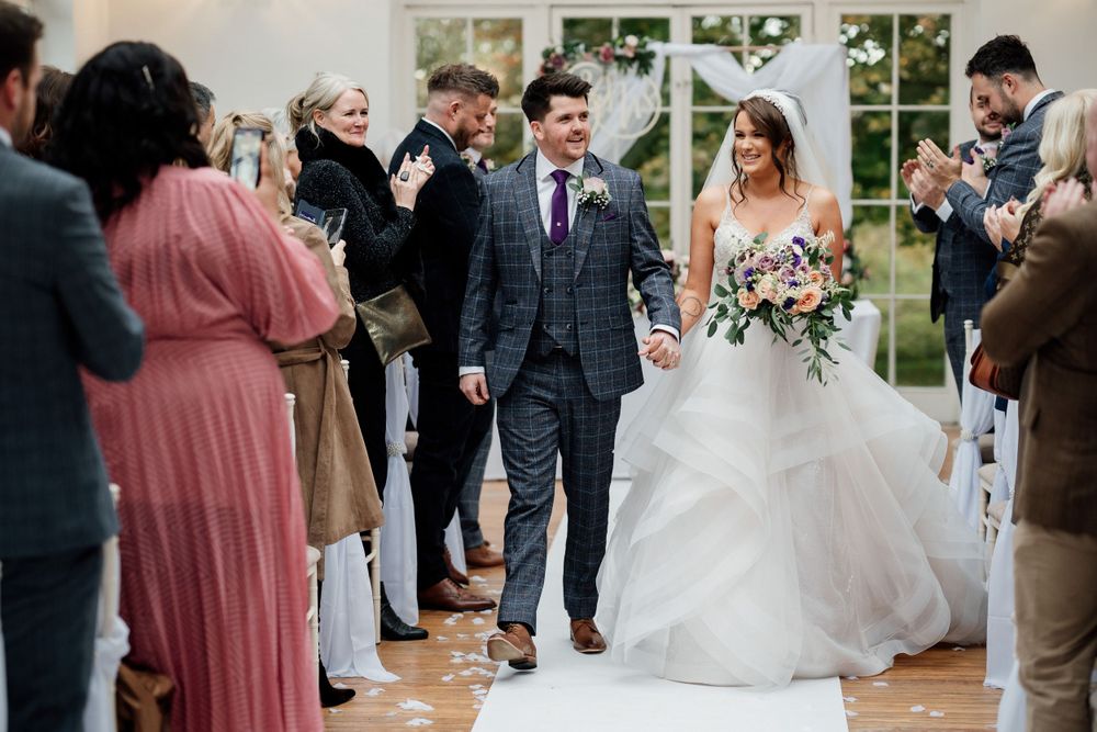 Newlyweds walking down the aisle while guests cheer in a light-filled ceremony space.