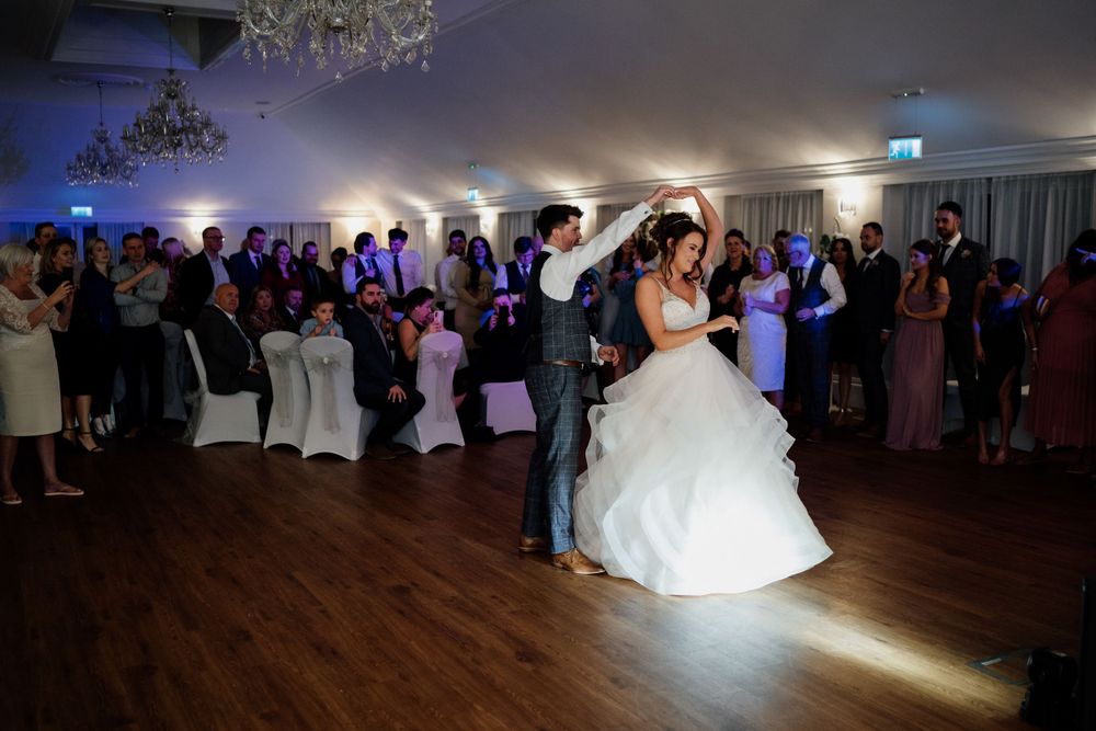 Newlyweds perform their first dance at Barton Hall under spotlights while guests watch in a ballroom setting.