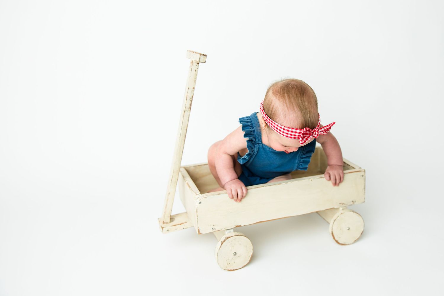 Small child in blue overalls sitting in a vintage white wooden wagon with wheels against white backdrop.