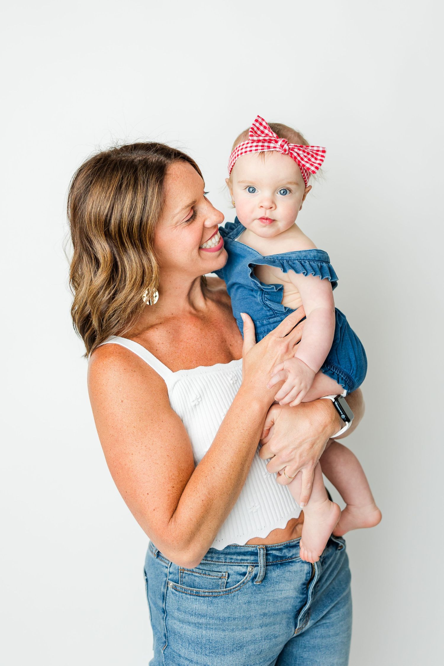 A tender moment showing a parent in white top and jeans holding a baby wearing denim and red gingham bow against white background.