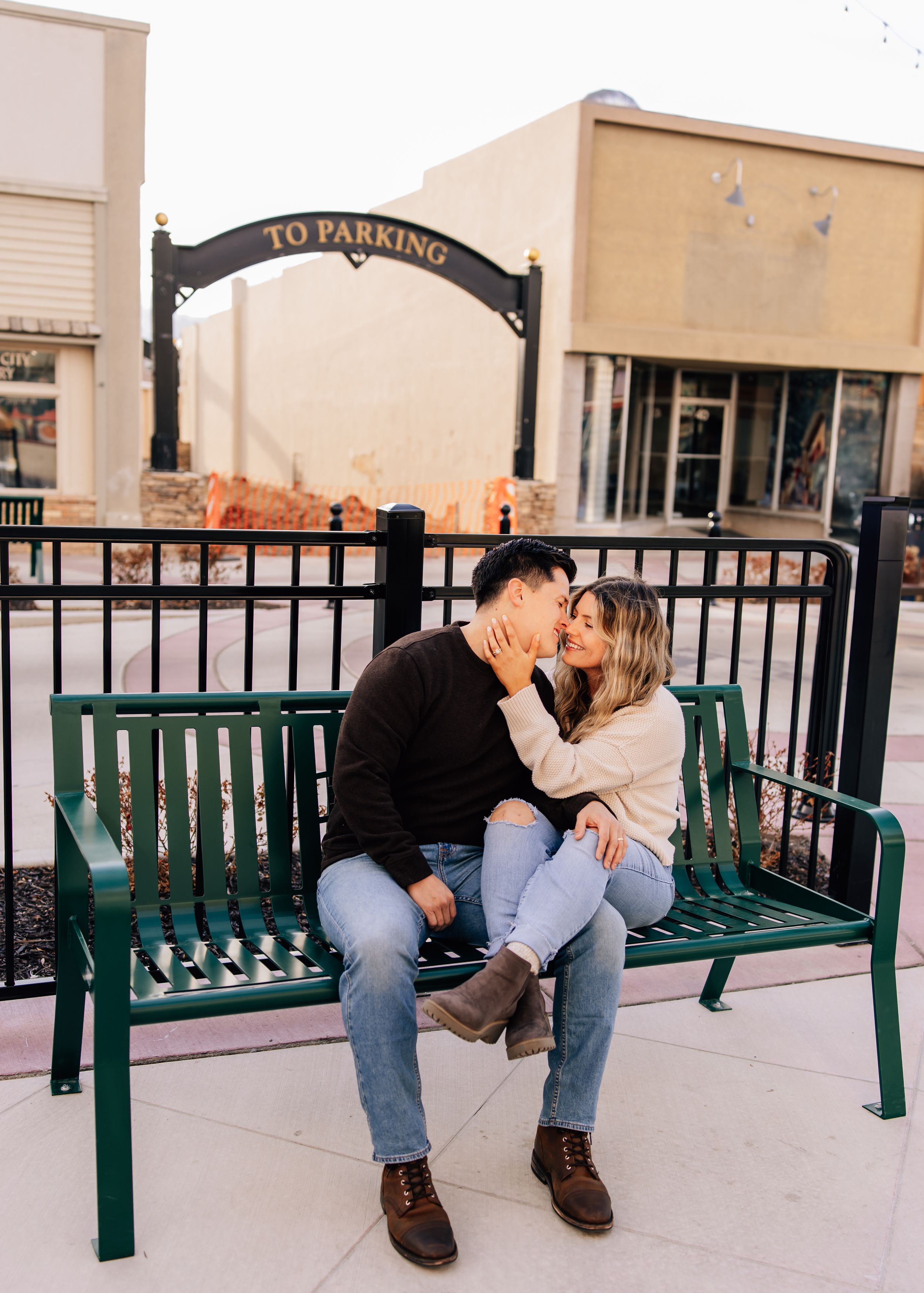 A couple shares a tender moment while sitting on a green park bench under an archway.