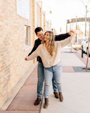 A playful couple spreads their arms while having fun on an urban sidewalk with brick buildings.
