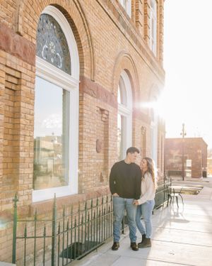 A couple leans against a historic brick building with arched windows during sunset.