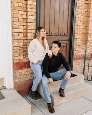 Couple in casual attire sitting on brick building steps during engagement photoshoot.