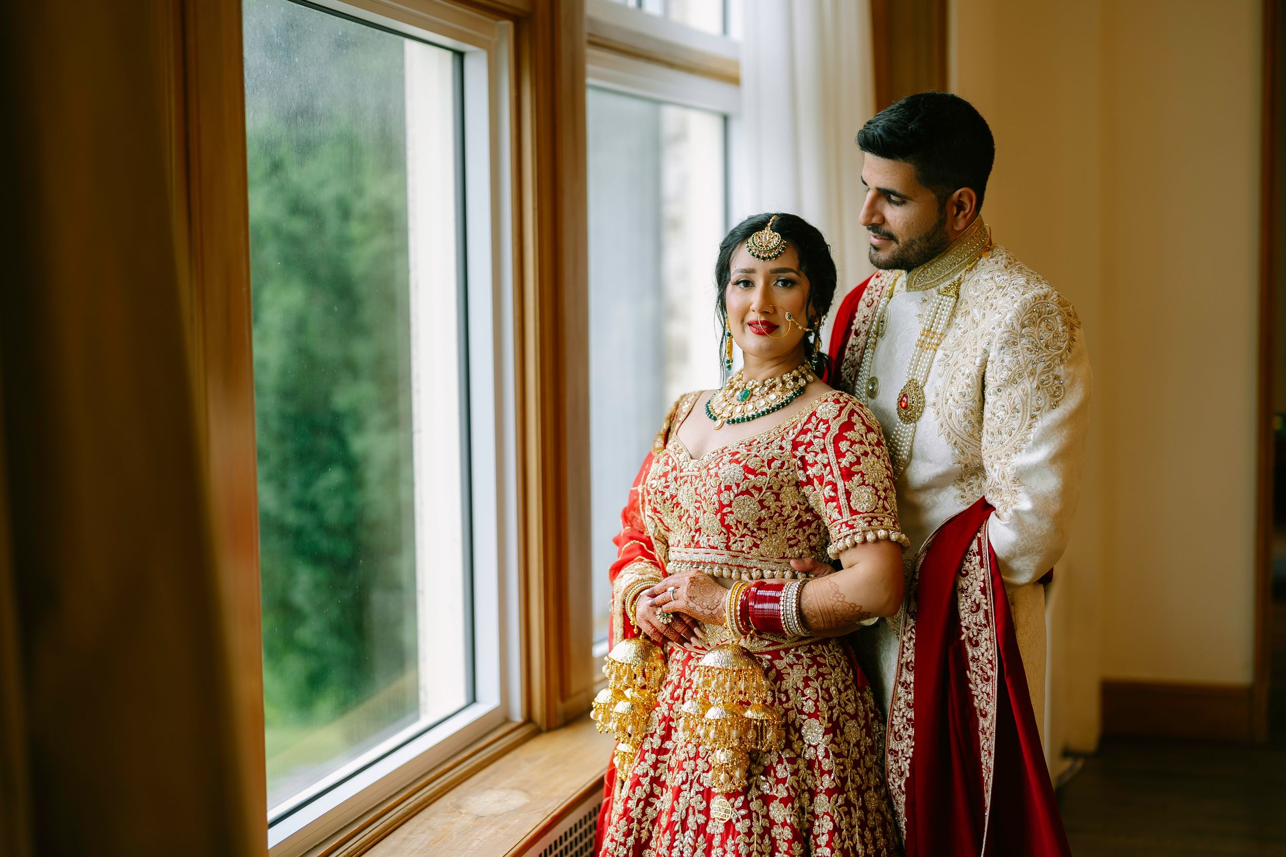 A couple in traditional South Asian wedding attire stands together by a window in warm indoor lighting.