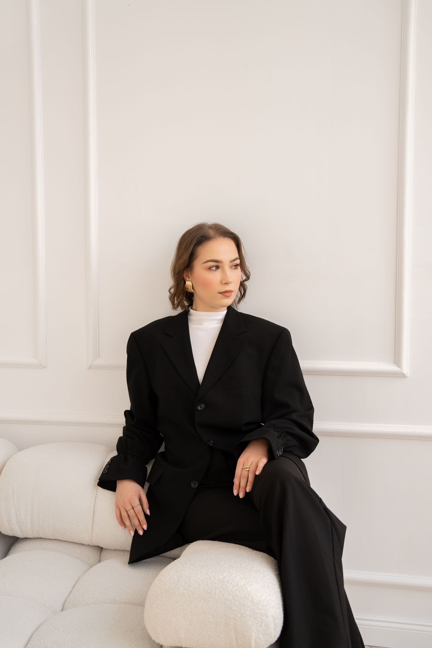 Professional model poses in oversized black blazer and white turtleneck on white couch in minimal setting.