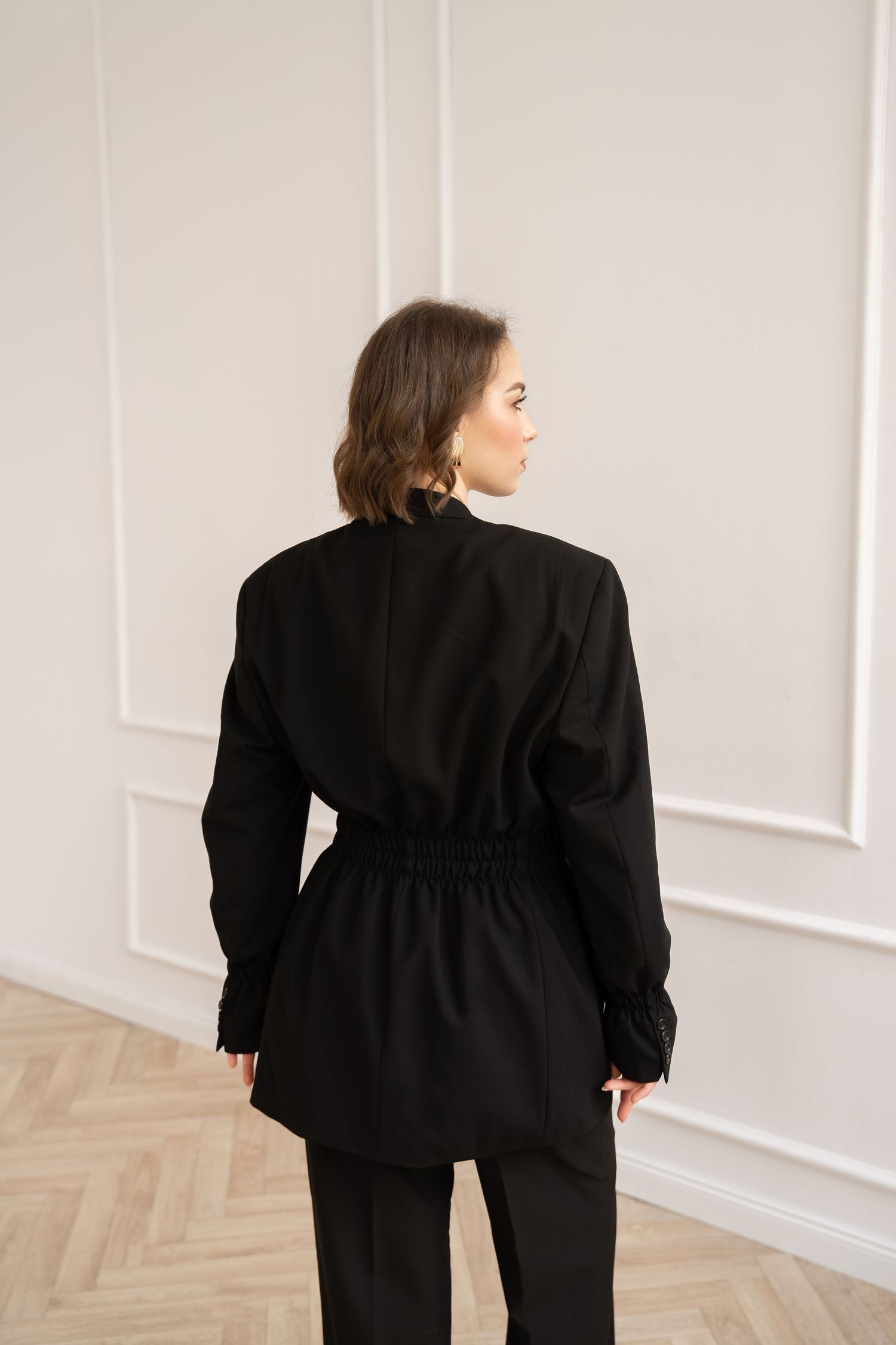 Back view of model wearing black tailored blazer with cinched waist against white paneled wall.
