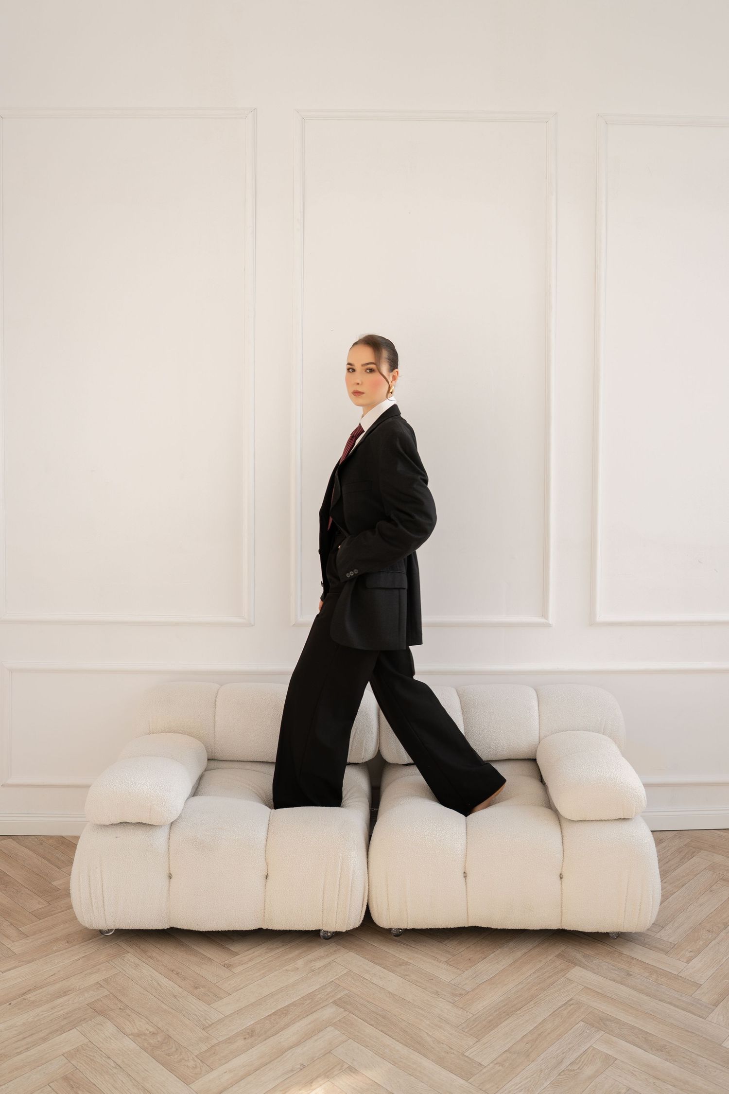 Model in black suit stands confidently on white modular sofa against minimalist wall.