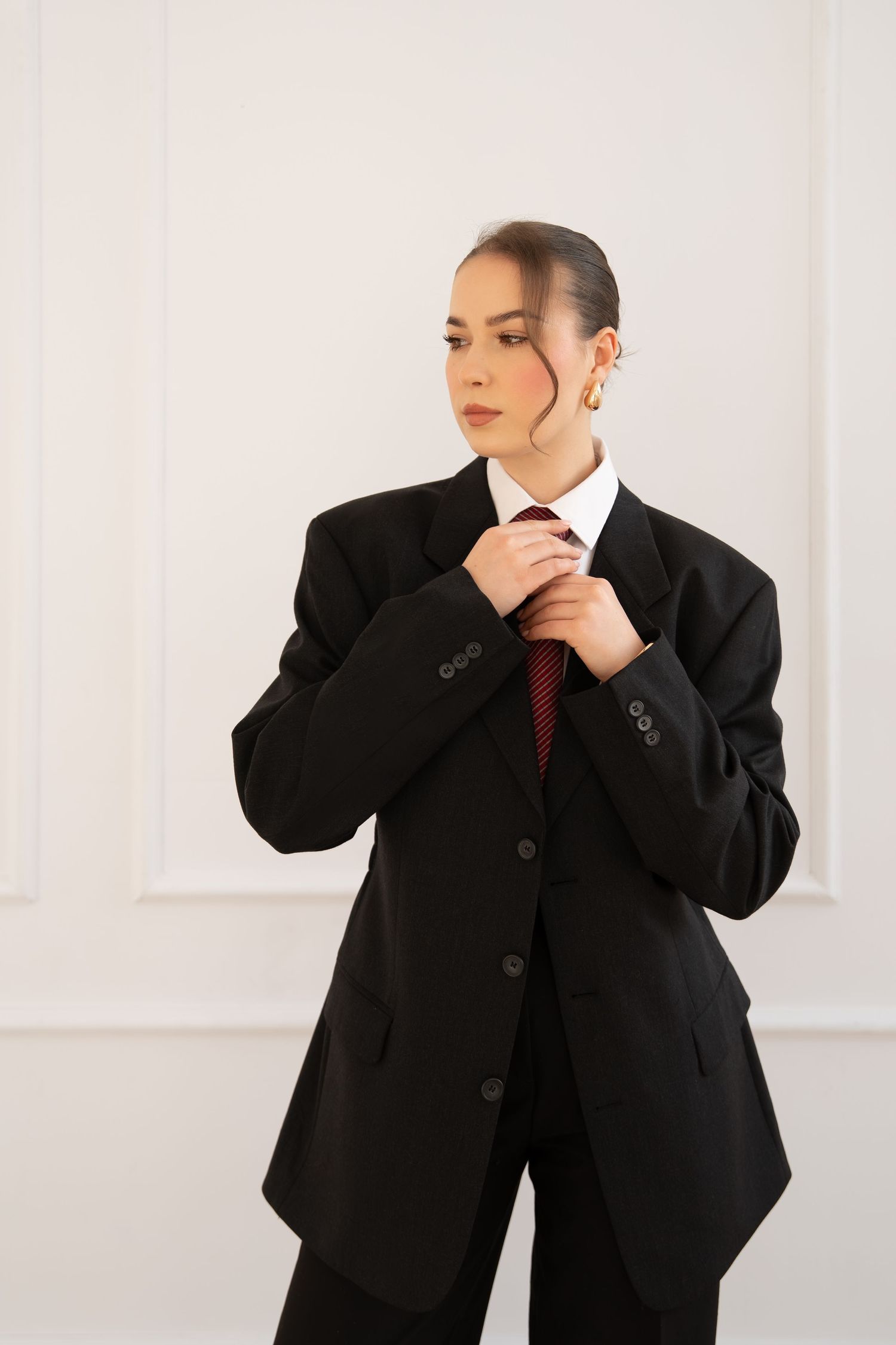 Fashion model adjusts tie while wearing oversized black suit jacket and white collar shirt.