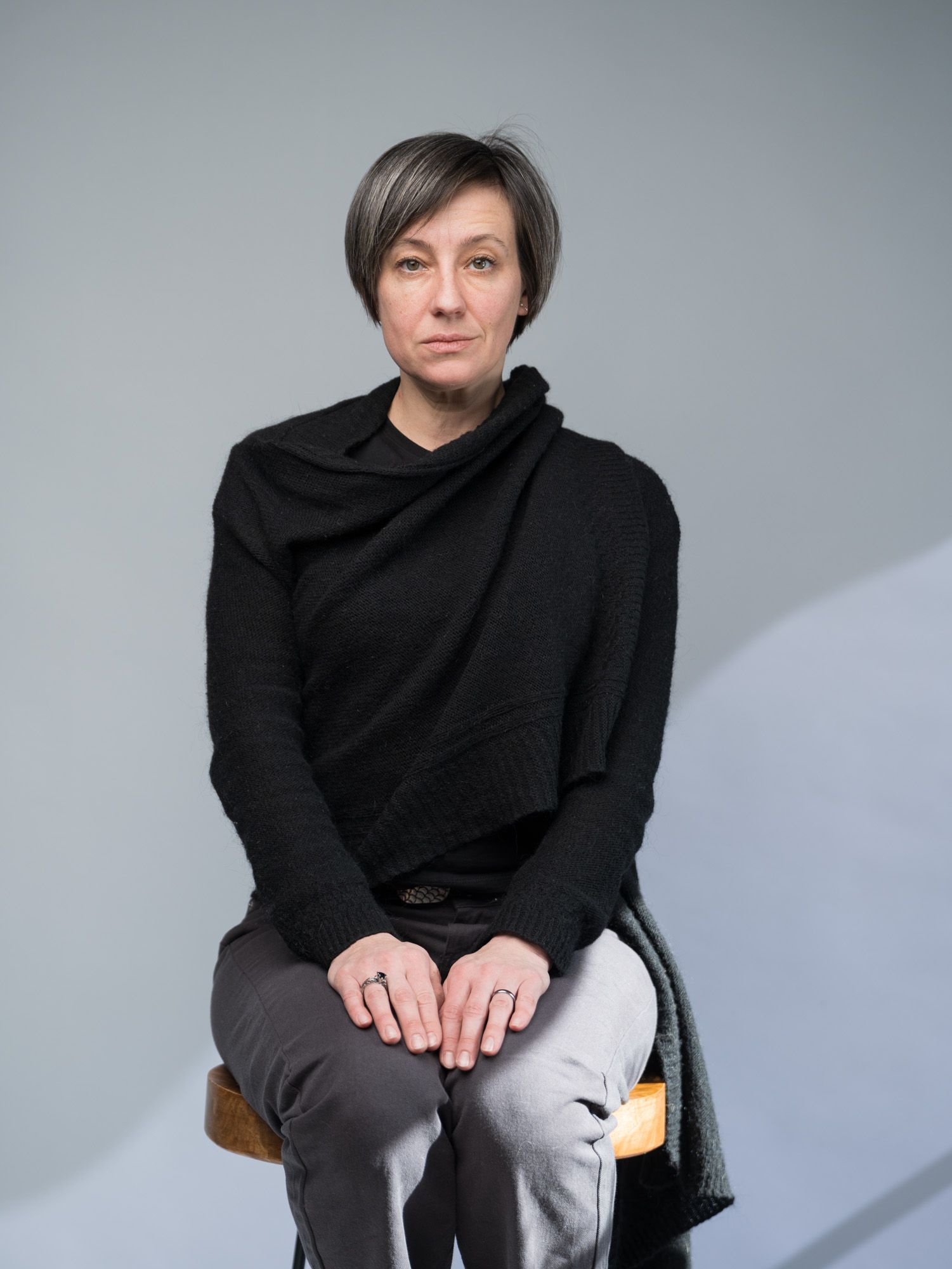 Figure in black sweater and gray pants sits in various poses against light studio backdrop.