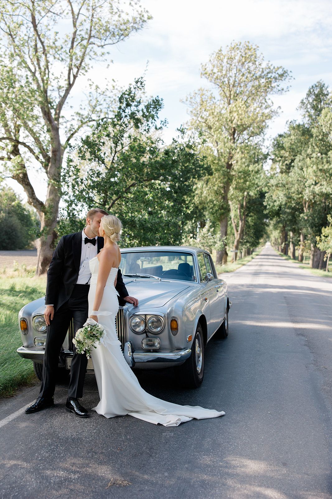 A couple poses with a classic vintage car on a tree-lined street during their wedding photoshoot.