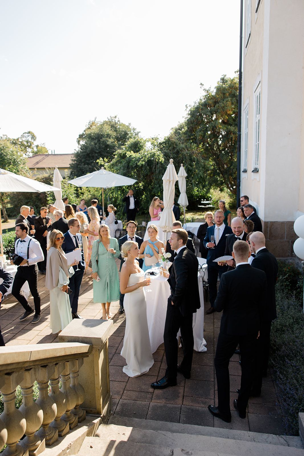 Wedding party in formal attire celebrates on an elegant outdoor terrace with white columns and stone balustrades.