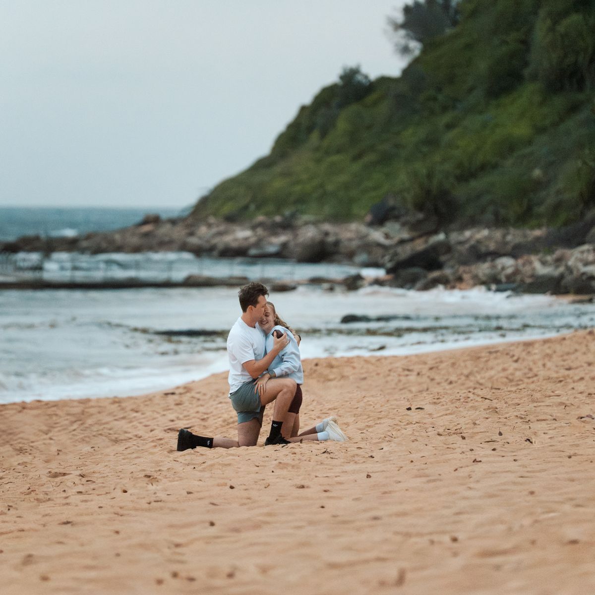 A person crouches on a sandy beach with rocky coastline and ocean waves in the background Newport photoshoot Sydney