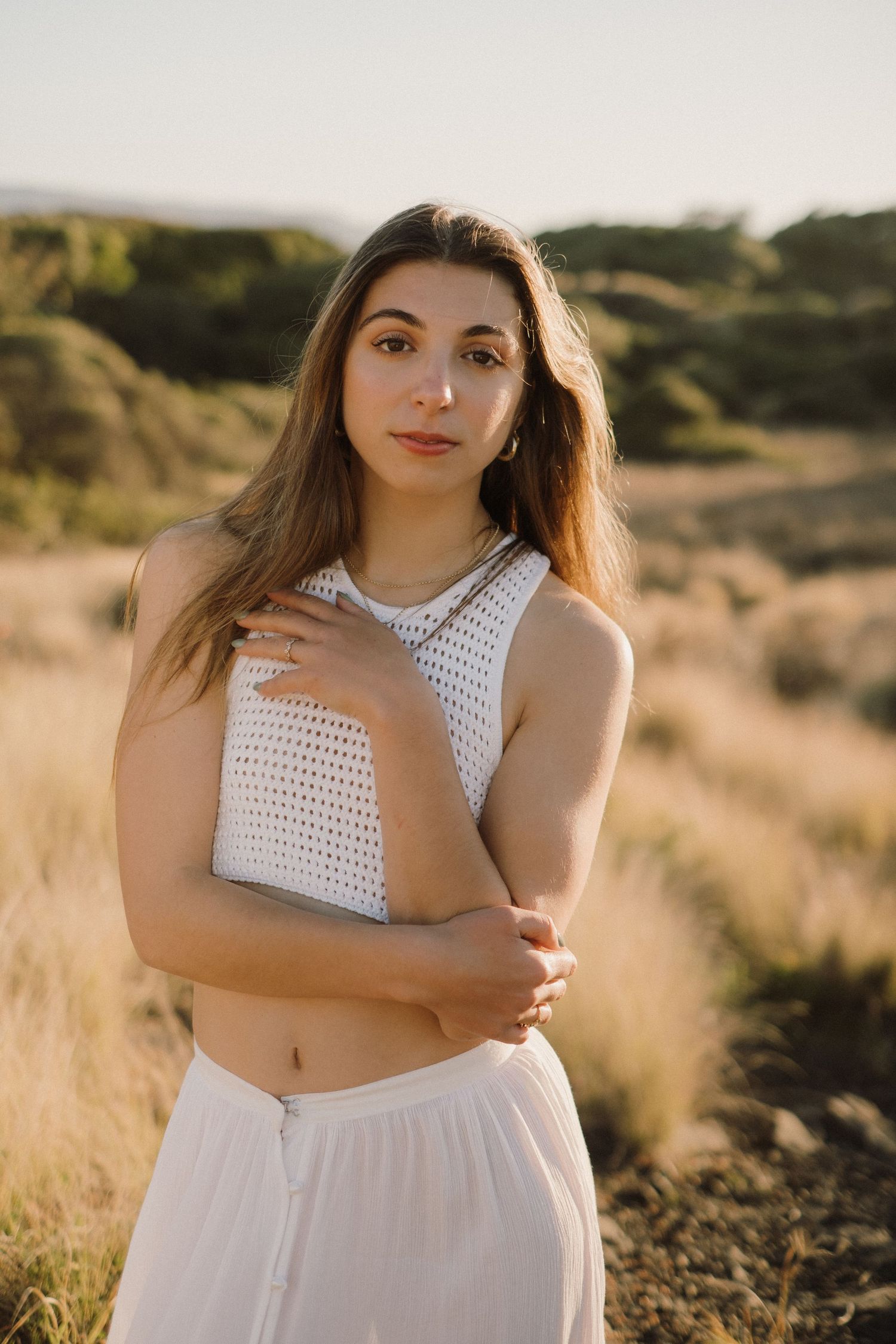 Woman in white crochet crop top poses outdoors during golden hour in natural setting.