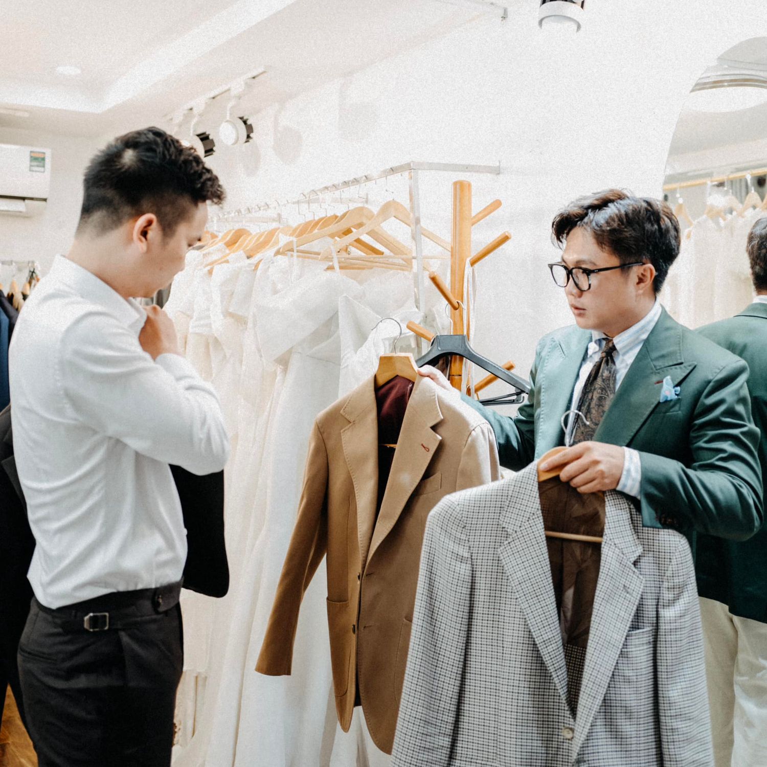 Two men browse suit jackets in an elegant bridal boutique with white gowns.