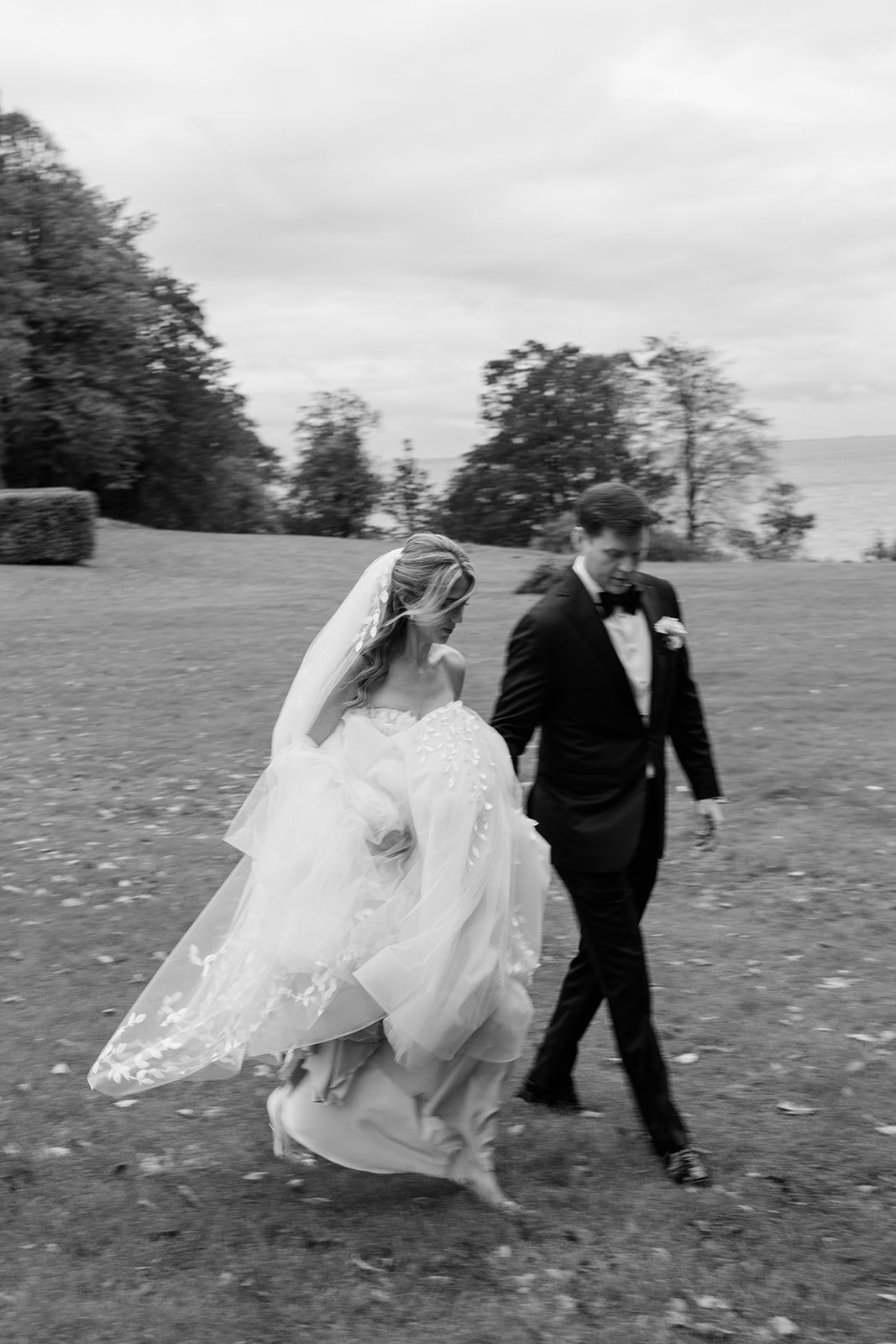 A couple in wedding attire runs playfully through a grassy field on an overcast day.
