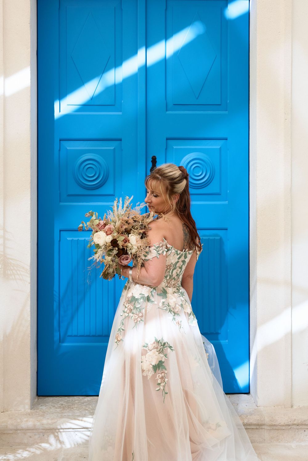 A bride in an embellished gown holds a dried flower bouquet against a bright blue door.