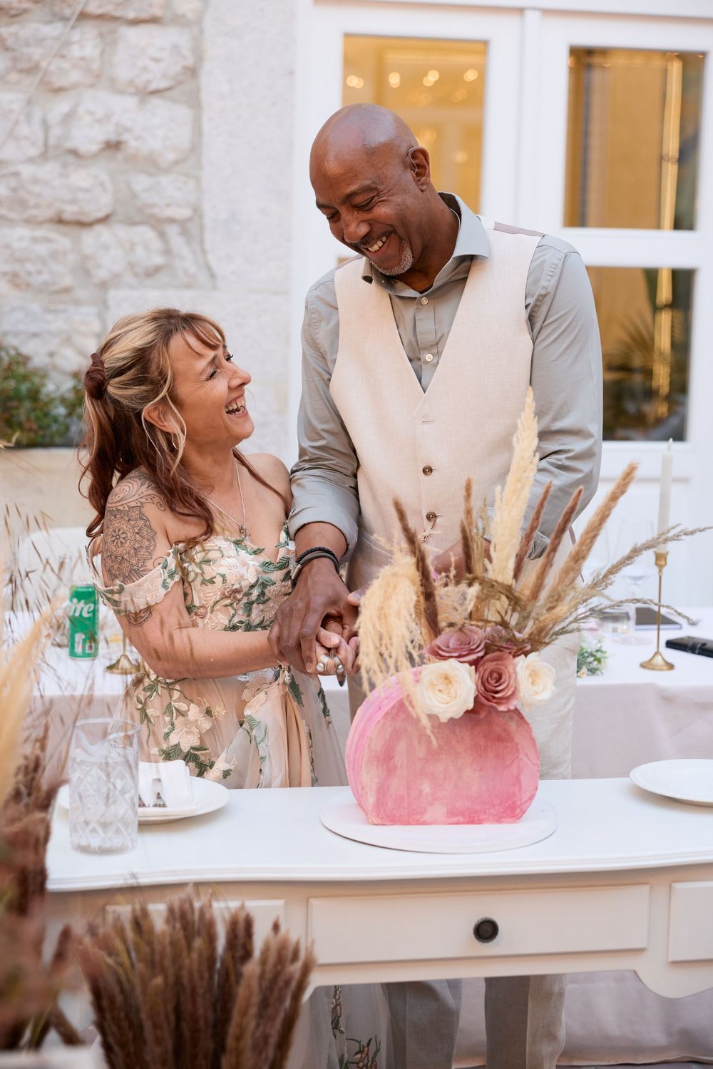 A joyful couple shares a laugh while celebrating with a pink cake at an elegant reception with string lights and stone walls.