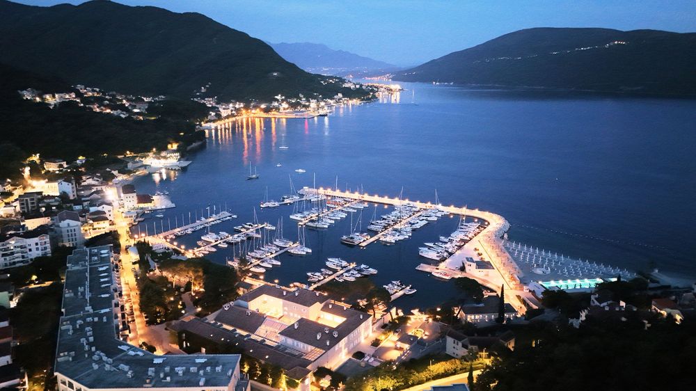 Panoramic nighttime view of a coastal marina with mountains, boats, and twinkling city lights reflecting on calm waters.