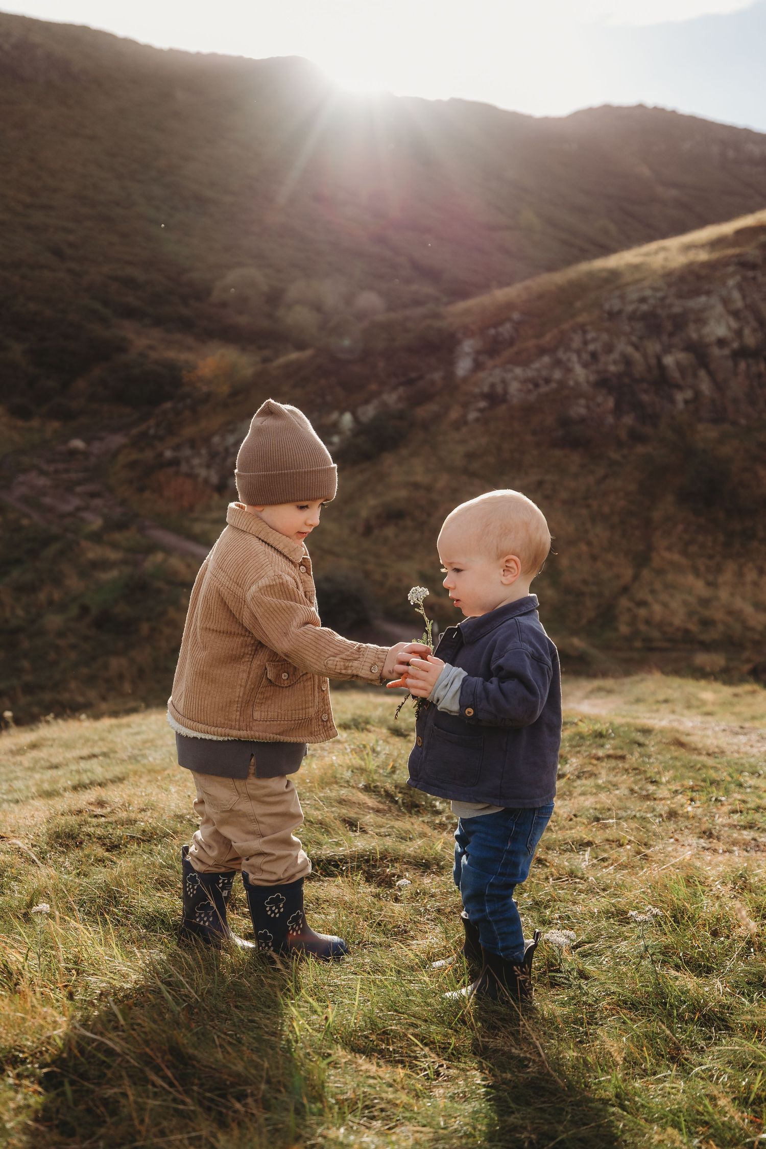 Two small children share dandelions together on a grassy hillside at sunset.