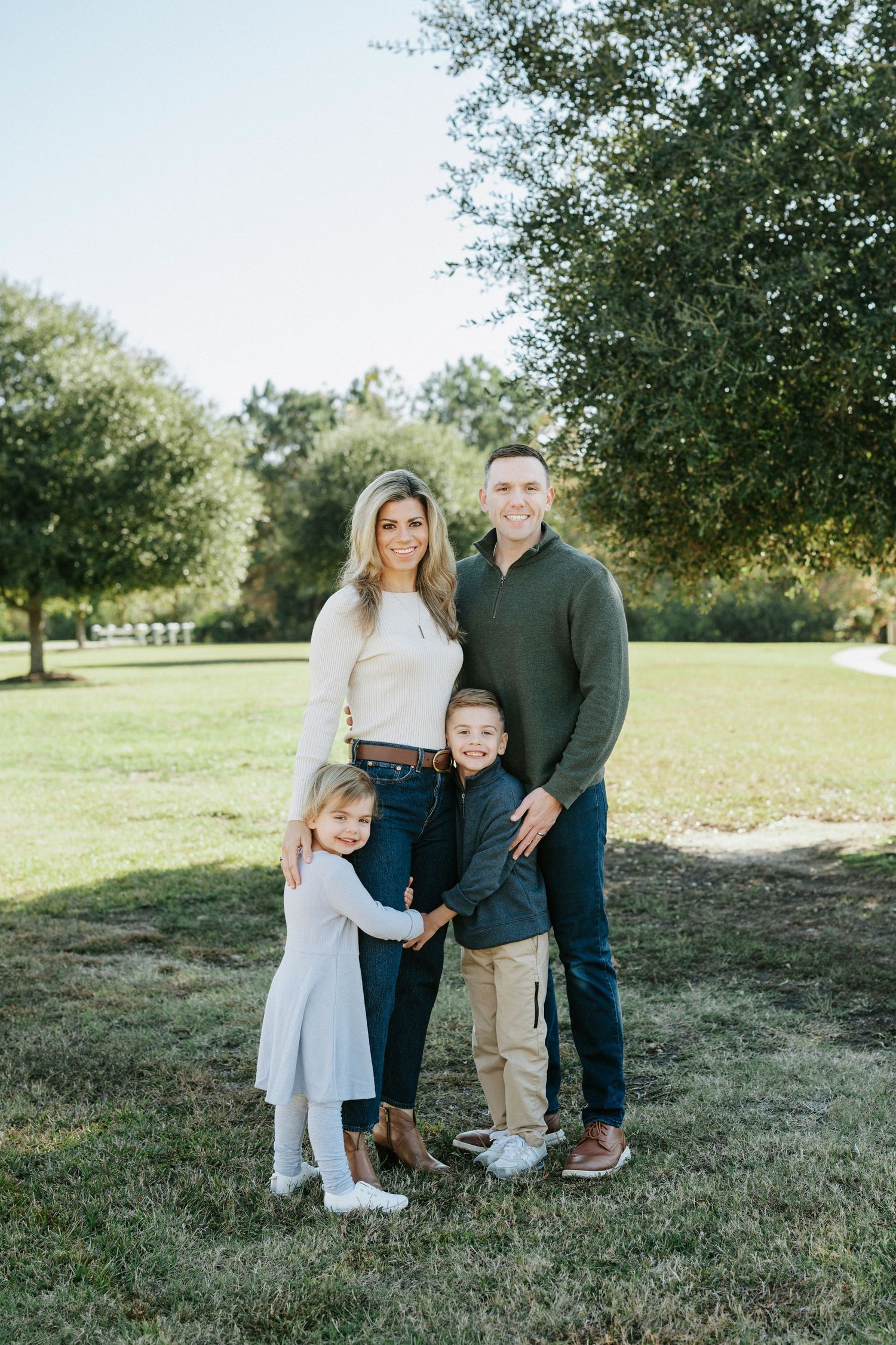 Family portrait session outdoors in a grassy field with trees in the background on a sunny day.