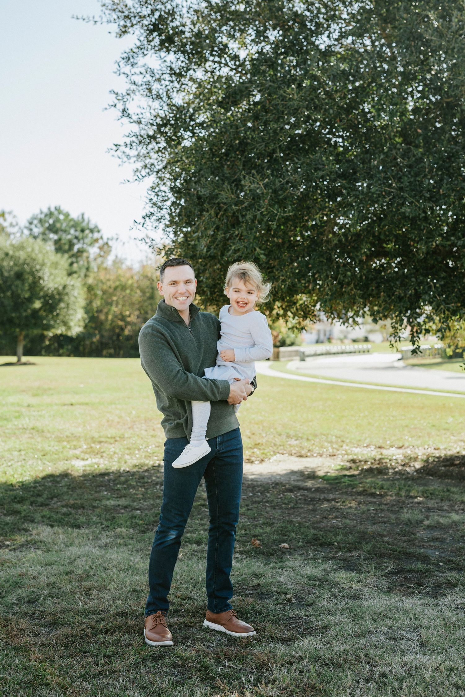 A couple embraces while standing in a grassy park area with trees in the background.