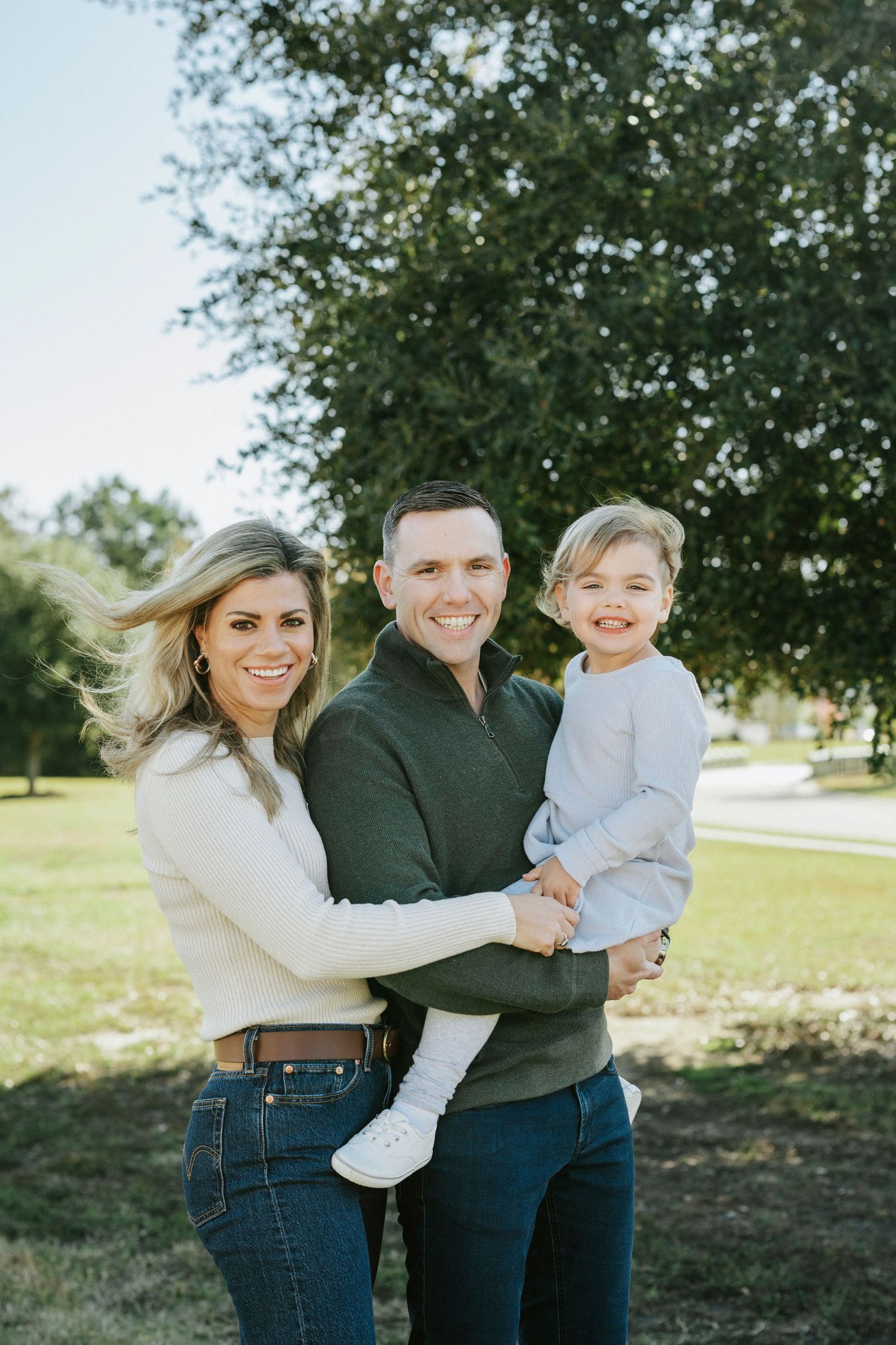 A family in casual attire embraces and laughs together while standing outdoors on a sunny day.