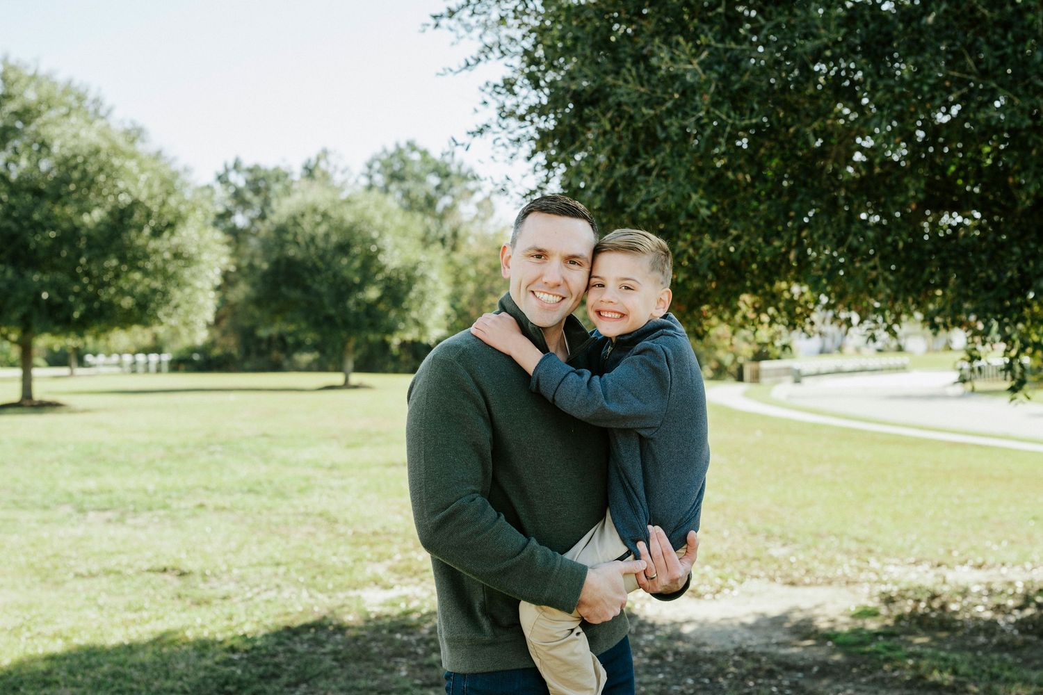 A couple shares tender moments together in a sunny park setting with green grass and trees in the background.