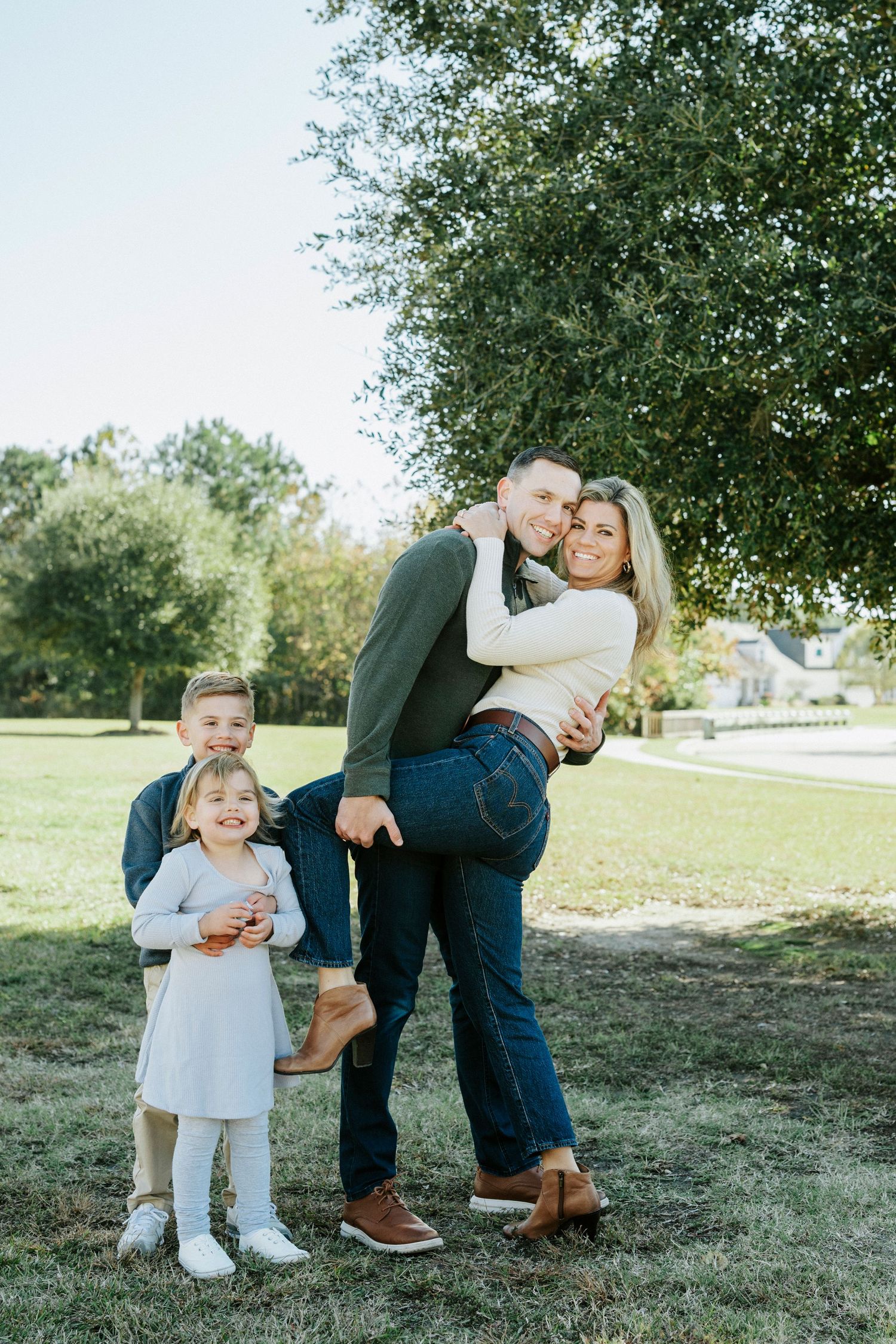 A young family embraces while standing together in a sunlit park setting during a casual photo shoot.