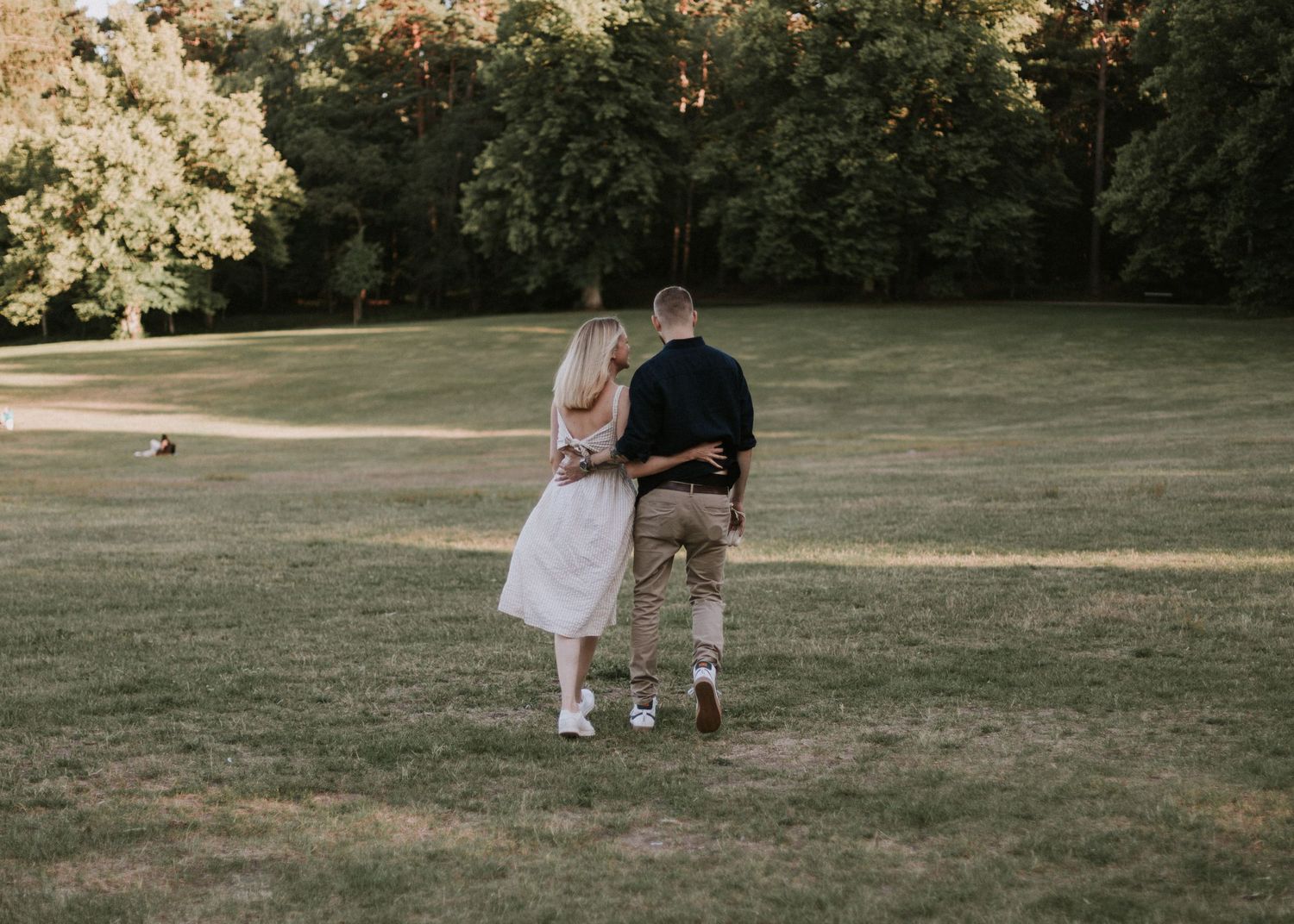 Couple walking arm in arm through sunny green park, woman in white dress and man in casual outfit.