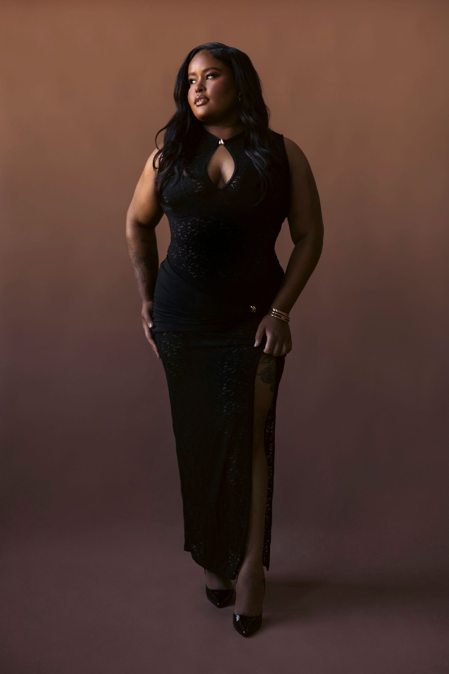 A figure in a flowing black evening dress poses elegantly against a dark brown studio backdrop.