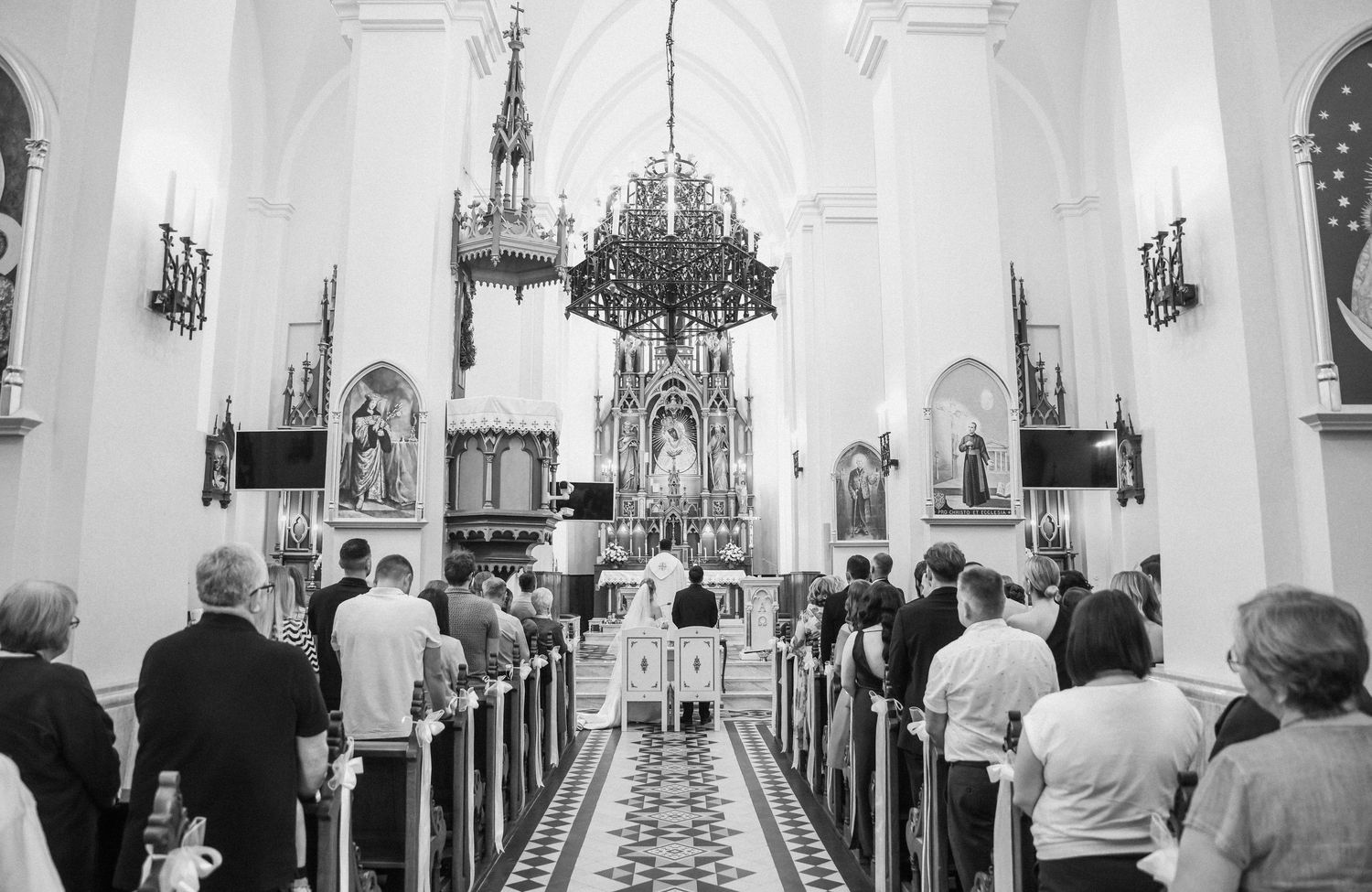 A black and white panoramic view of guests standing inside a church sanctuary with ornate chandeliers and pews.