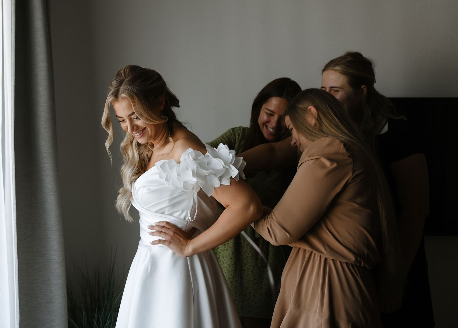 A group of women help adjust a white wedding dress in a dimly lit room with curtains.