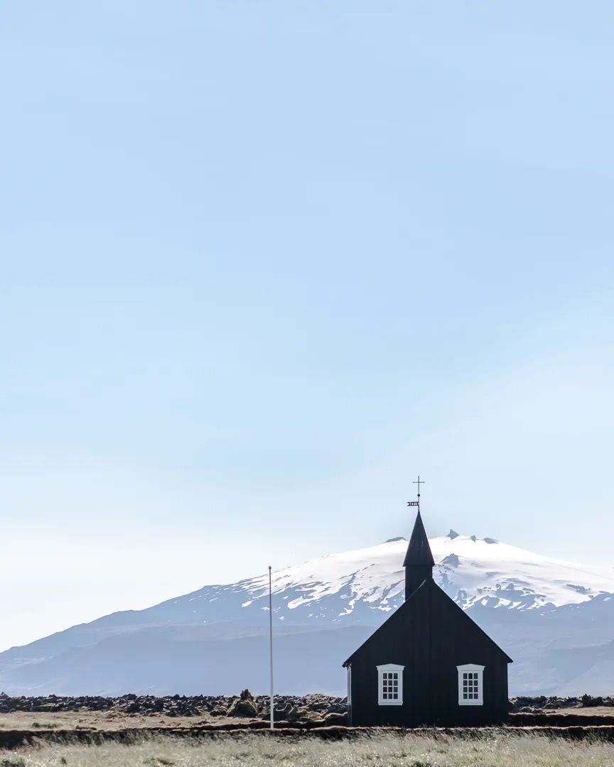 Black wooden church standing alone with snow-topped mountains behind it.