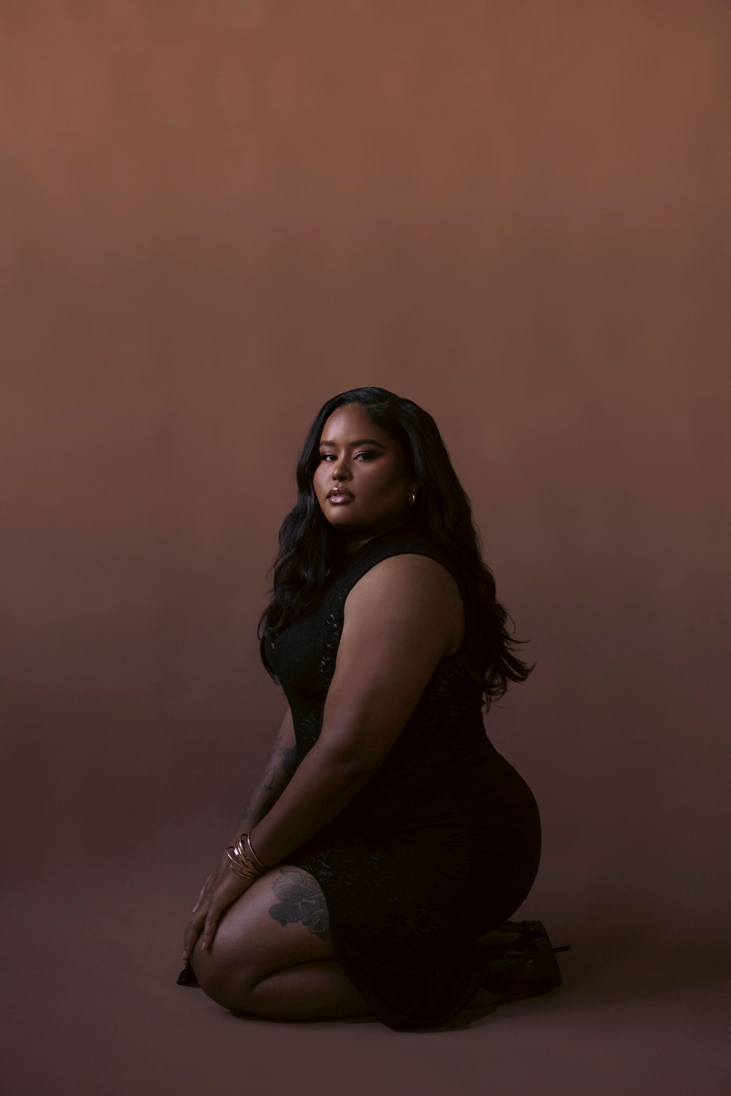 A figure in a black dress poses elegantly against a dark brown backdrop in a moody artistic portrait.