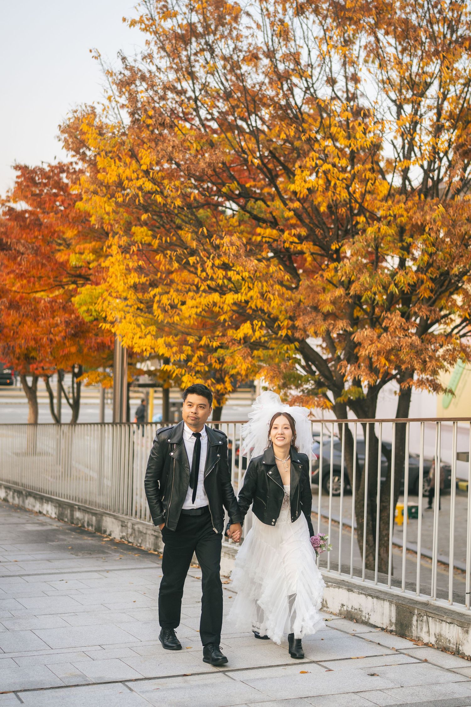 A couple walks hand in hand along a sidewalk beneath vibrant orange and yellow autumn trees.