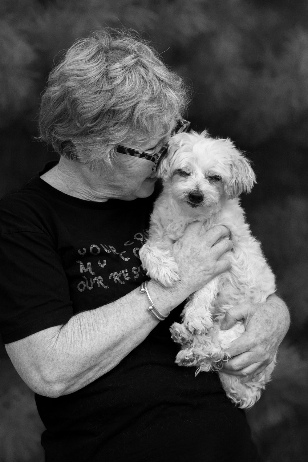 A small white fluffy dog being held and cuddled by someone wearing a black t-shirt in a black and white photo.