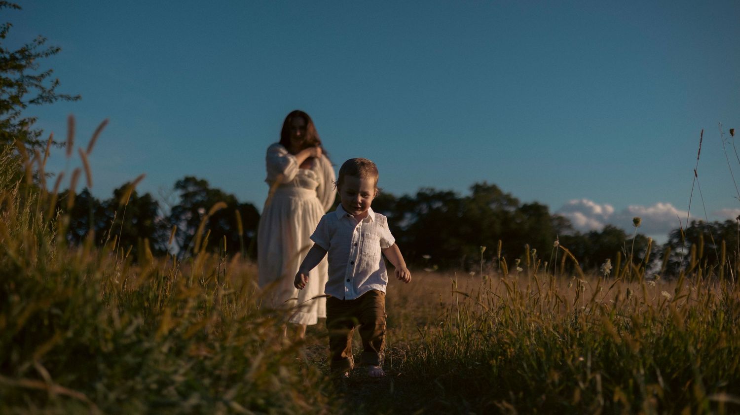 A couple in white attire walks through a golden wheat field during sunset creating a romantic silhouette.