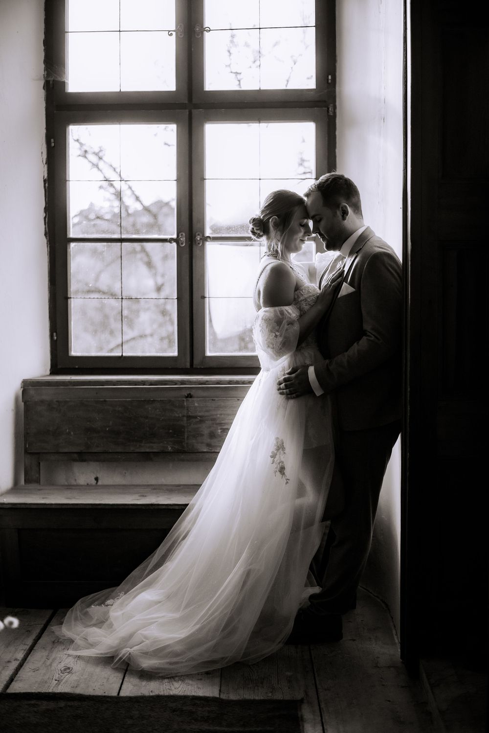 Dramatic black and white wedding photo of couple embracing by vintage windows.