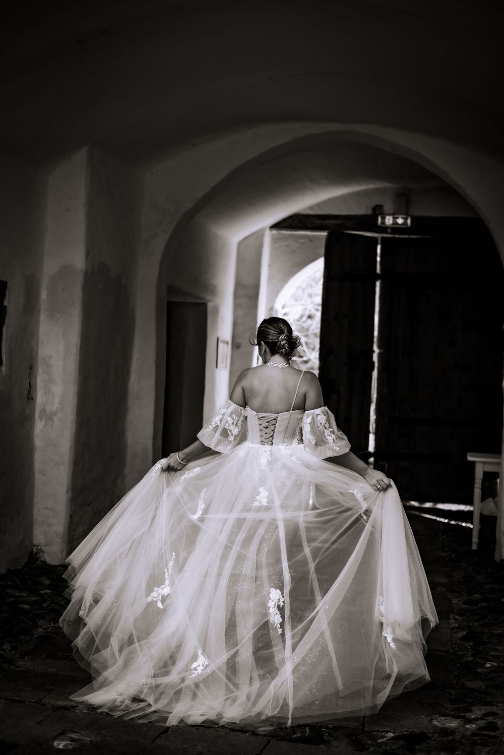 Artistic black and white photo of flowing wedding dress in an arched hallway.